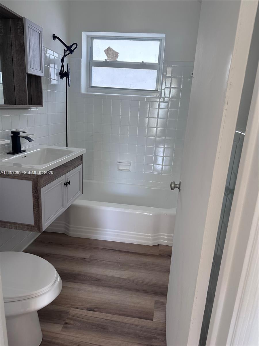 Bathroom featuring wood-finish flooring, a white porcelain tub with integrated tile surround, wall-mounted medicine cabinet, and vanity with a matte black faucet