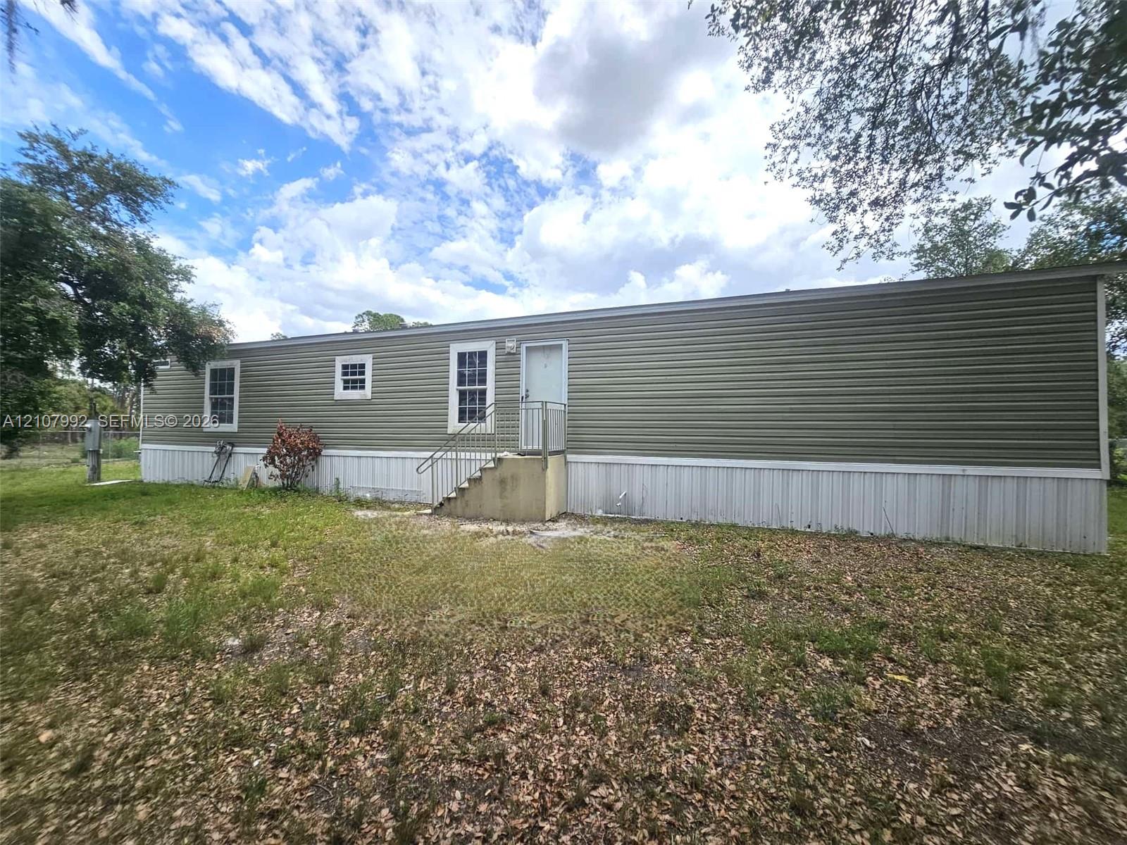 Olive green vinyl siding exterior featuring white metal skirting, three sash windows, and concrete stairs with a metal handrail