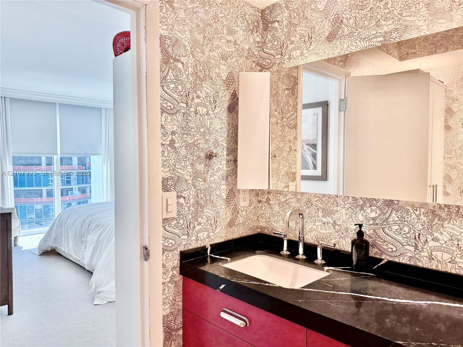 Bathroom featuring a deep-toned vanity cabinet, integrated dark stone-finish countertop with a rectangular undermount sink, chrome widespread faucet, and large-scale wall mirror set against patterned wallpaper