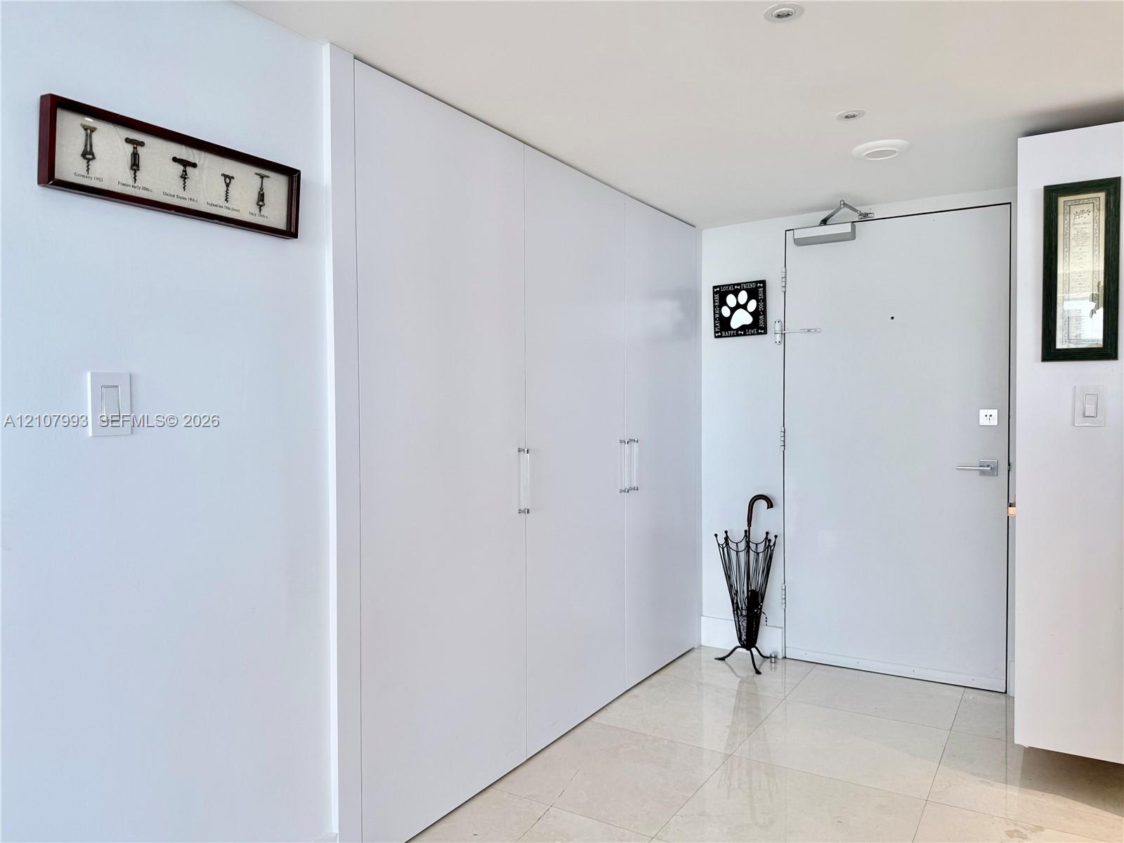 Floor-to-ceiling built-in white cabinetry with chrome hardware, polished stone tile flooring, and solid-core metal entry door with overhead hydraulic closer
