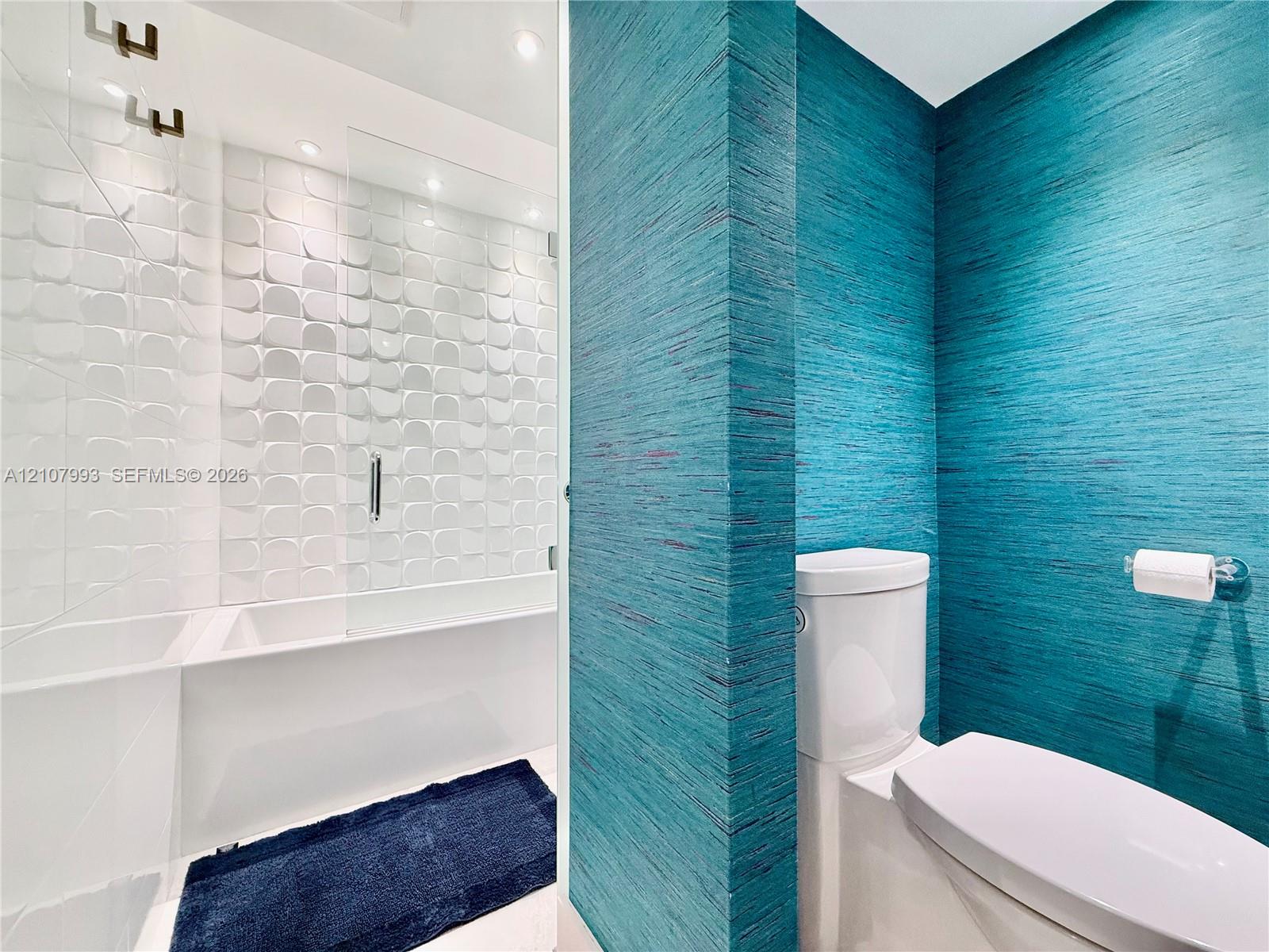Textured teal wall covering, geometric white tiled shower surround, glass shower partition, and white dual-flush toilet