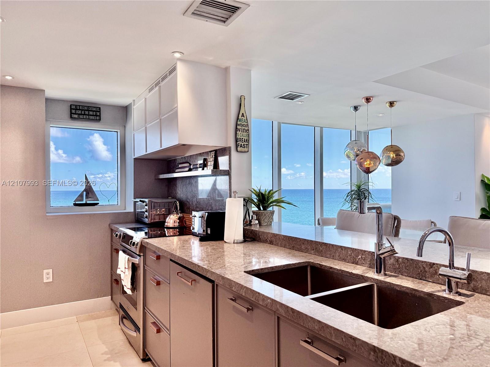 Kitchen featuring flat-panel cabinetry, stone countertops, stainless steel appliances, double-basin undermount sink with high-arc faucet, and floor-to-ceiling windows with coastal scenery