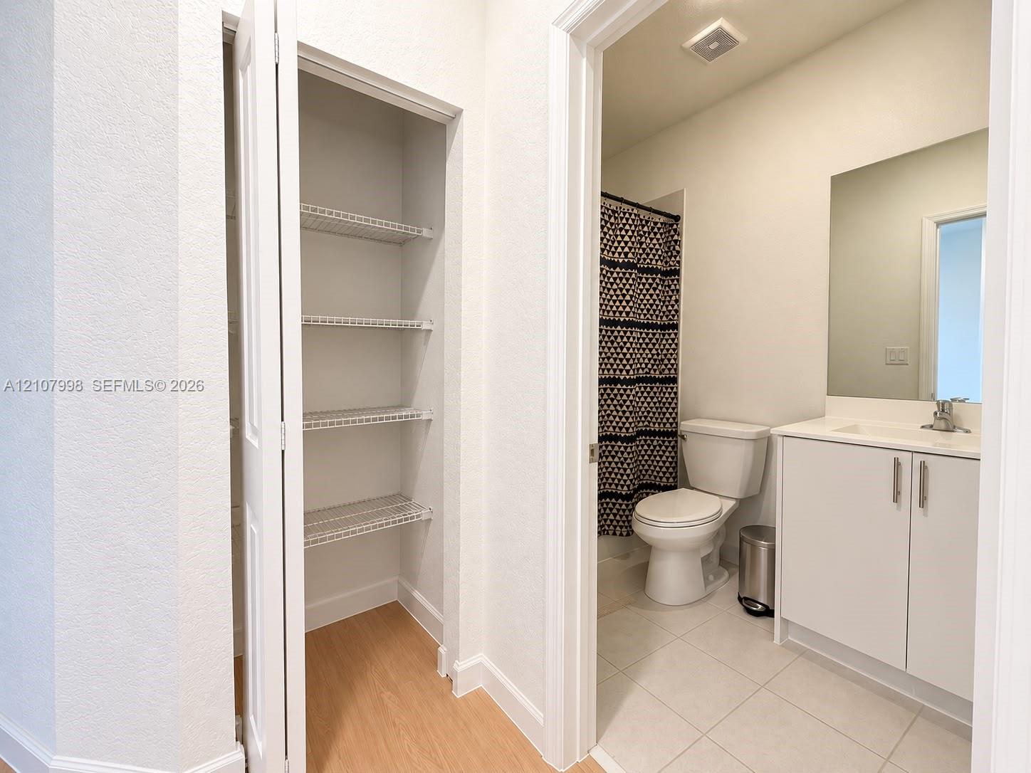 Built-in closet with wire shelving, white vanity cabinet with integrated sink, rectangular frameless mirror, and neutral tile flooring