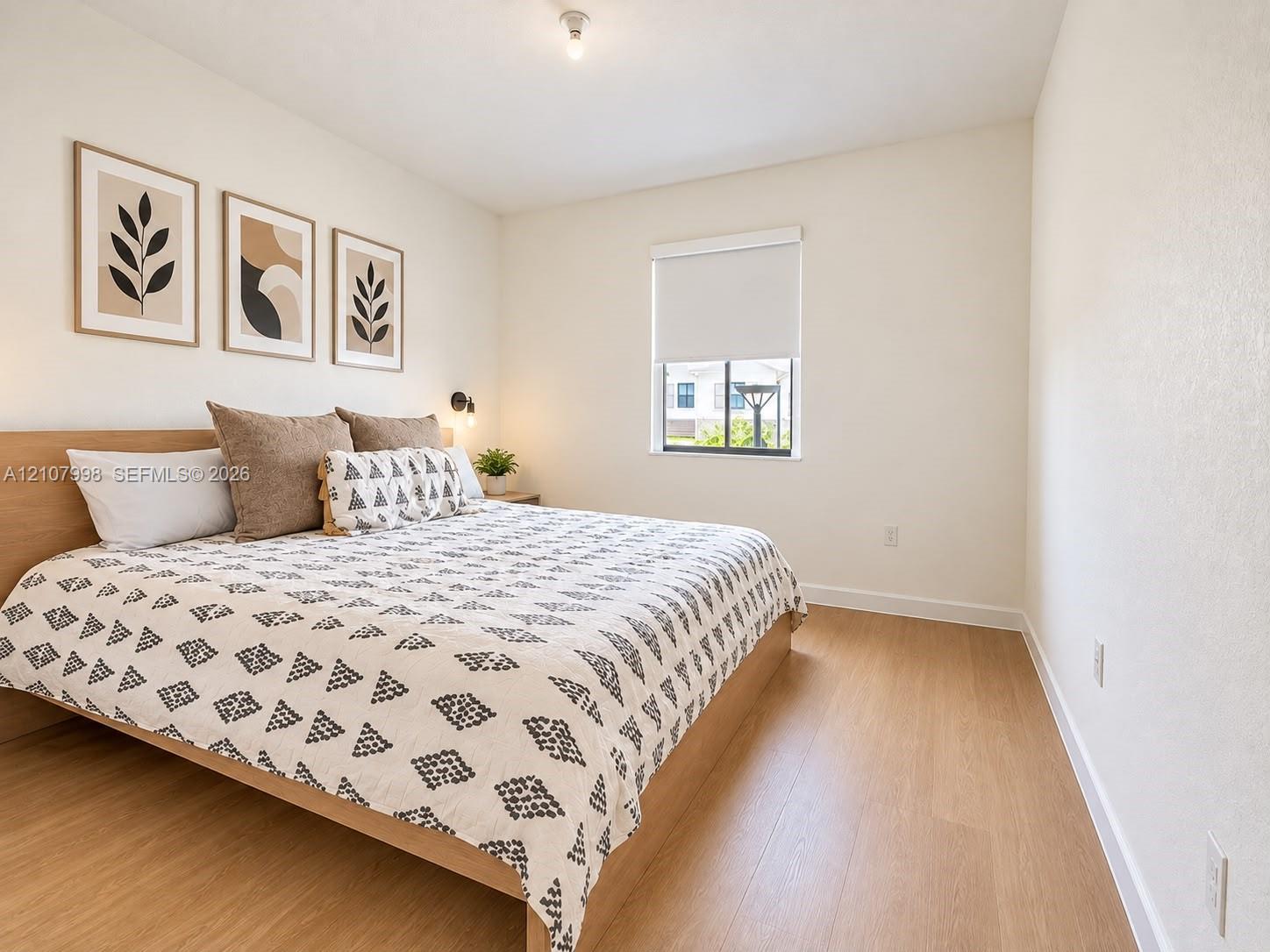 Neutral-toned bedroom featuring wood-finish flooring, a single-pane window with a roller shade, and a built-in wall-mounted light fixture