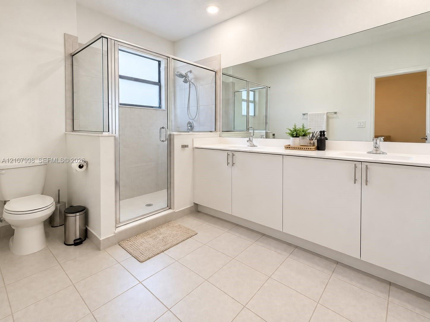 Dual-sink vanity with white cabinetry and solid surface countertop, large wall-mounted mirror, glass-enclosed shower with tile surround, and tile flooring