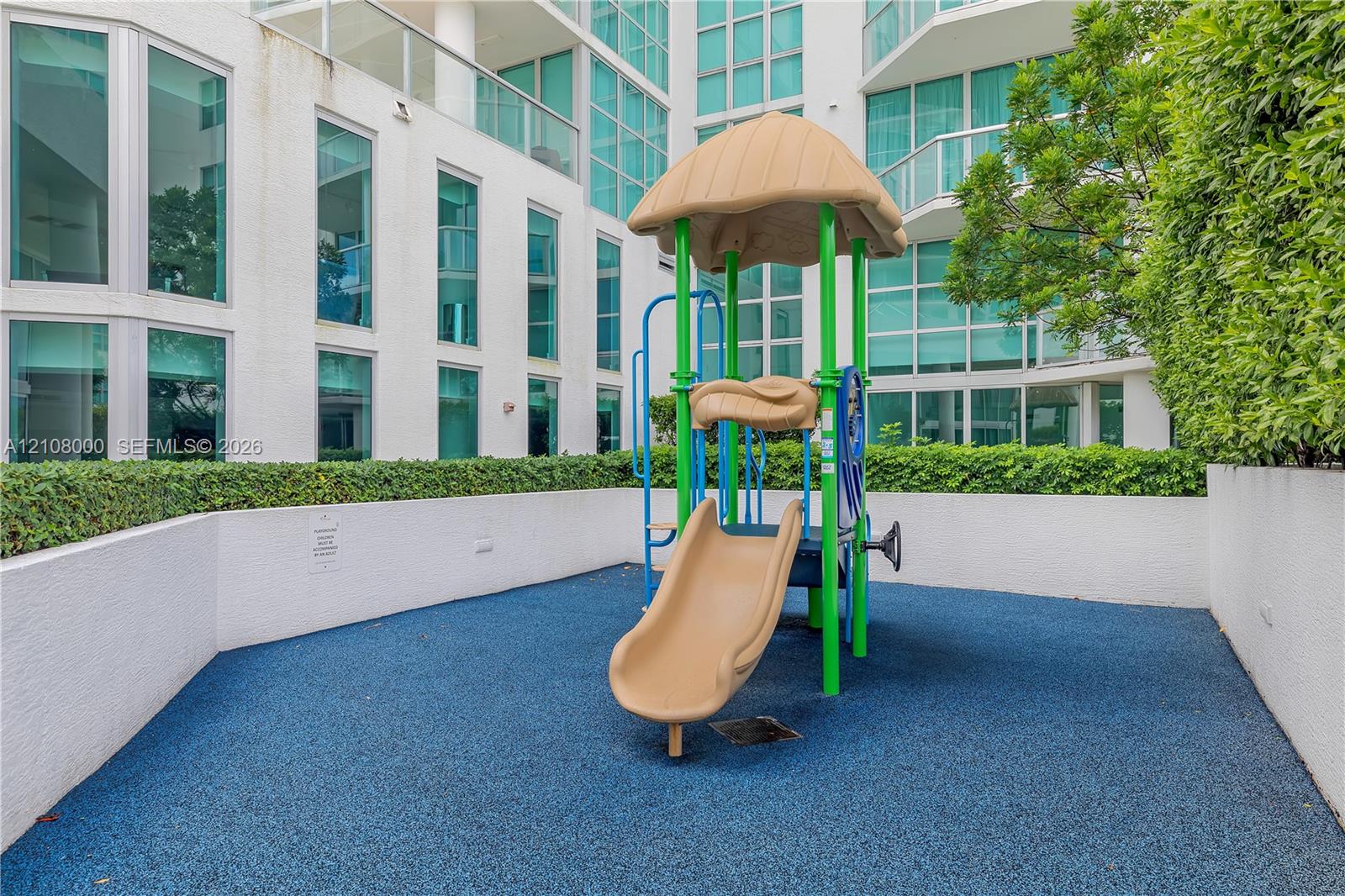 Outdoor recreational area featuring a structured play system with a slide, resilient rubberized flooring, and perimeter privacy hedges