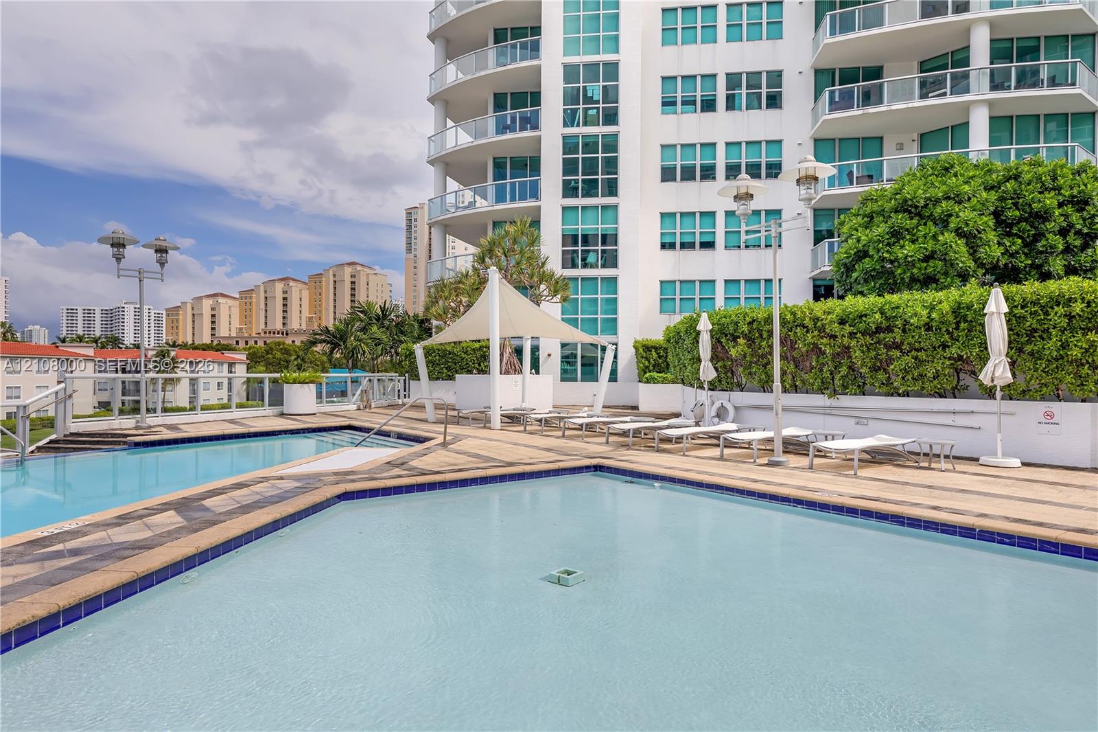 Multi-level outdoor pool deck featuring tile surround, shaded seating area with canopy, and perimeter landscaping