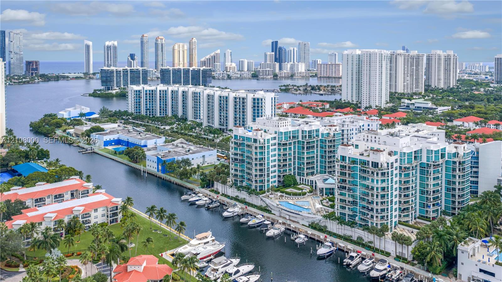 Aerial perspective of a multi-story residential complex featuring waterfront access, private boat slips, extensive hardscaping, and a swimming pool, situated against an urban skyline backdrop