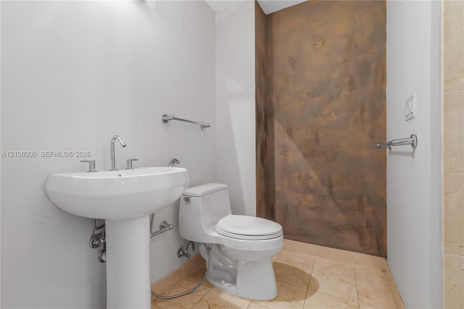 White porcelain pedestal sink with polished chrome faucet, dual-flush toilet, stone-finish tile flooring, and metallic-finish accent wall