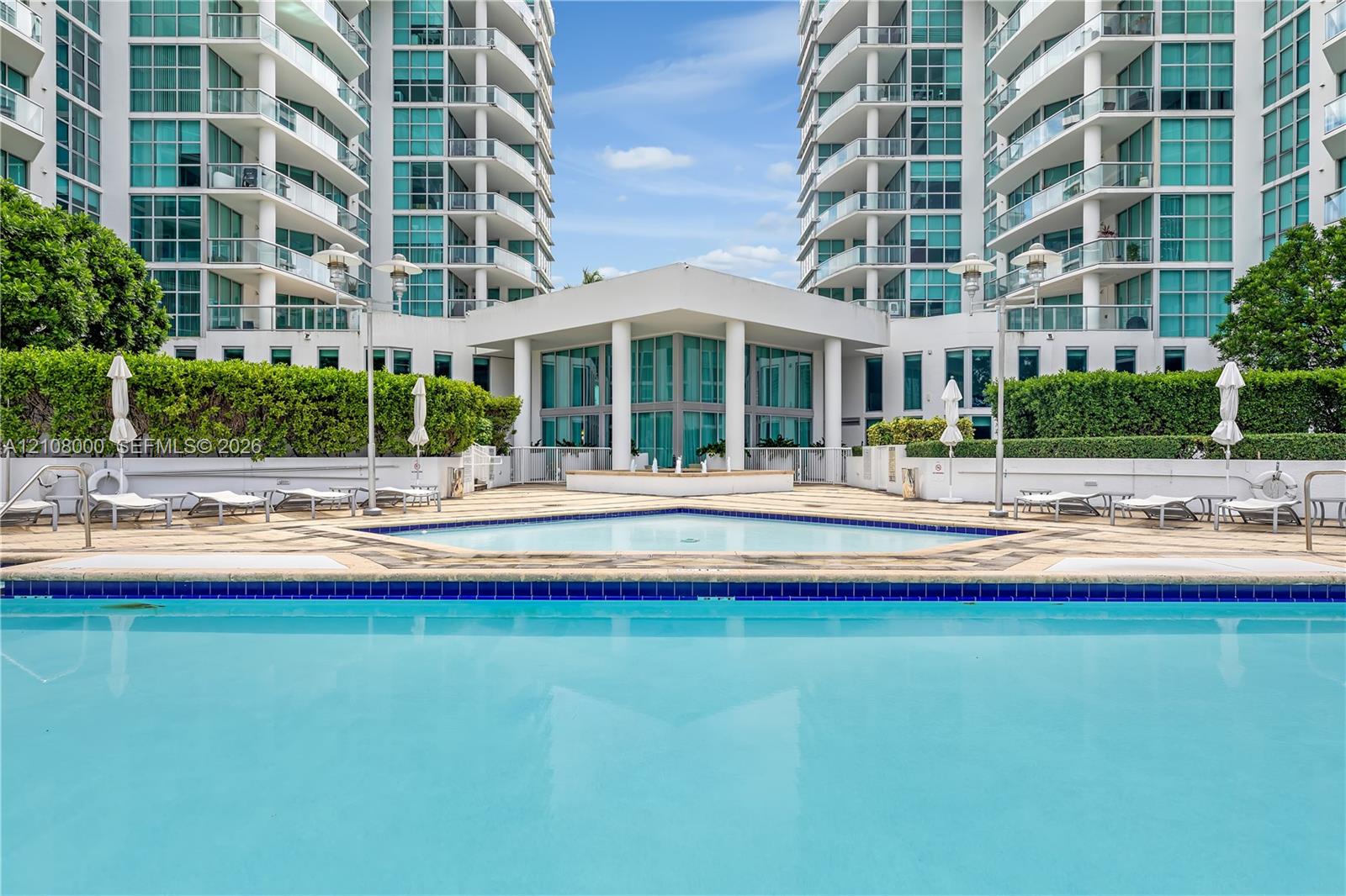 Swimming pool with decorative tile border, expansive stone-paver sun deck, manicured hedges, and multi-story high-rise architecture with glass-paneled balconies