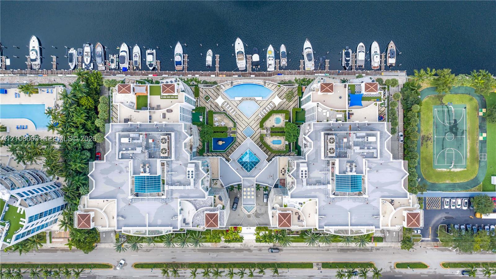 Aerial perspective of a multi-structure property featuring deep-water boat slips, multiple swimming pools, a hard-court sports facility, and landscaped grounds