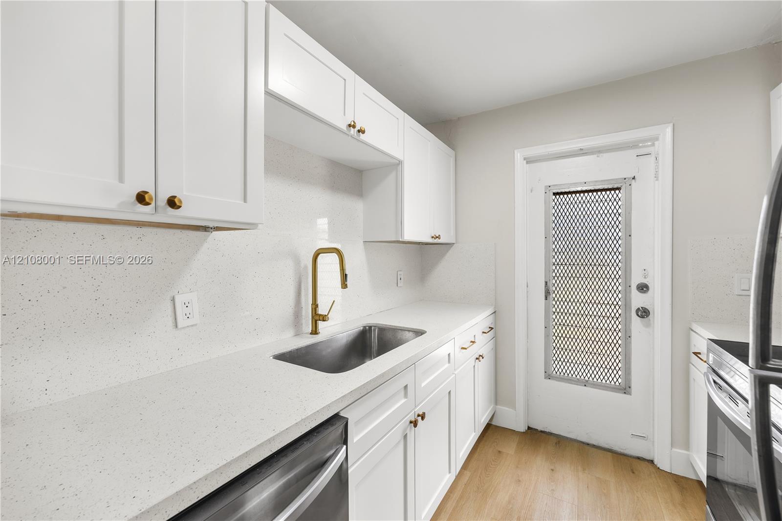 Kitchen featuring white shaker-style cabinetry, solid-surface countertops, a stainless steel undermount sink with a gold-tone gooseneck faucet, and wood-finish flooring