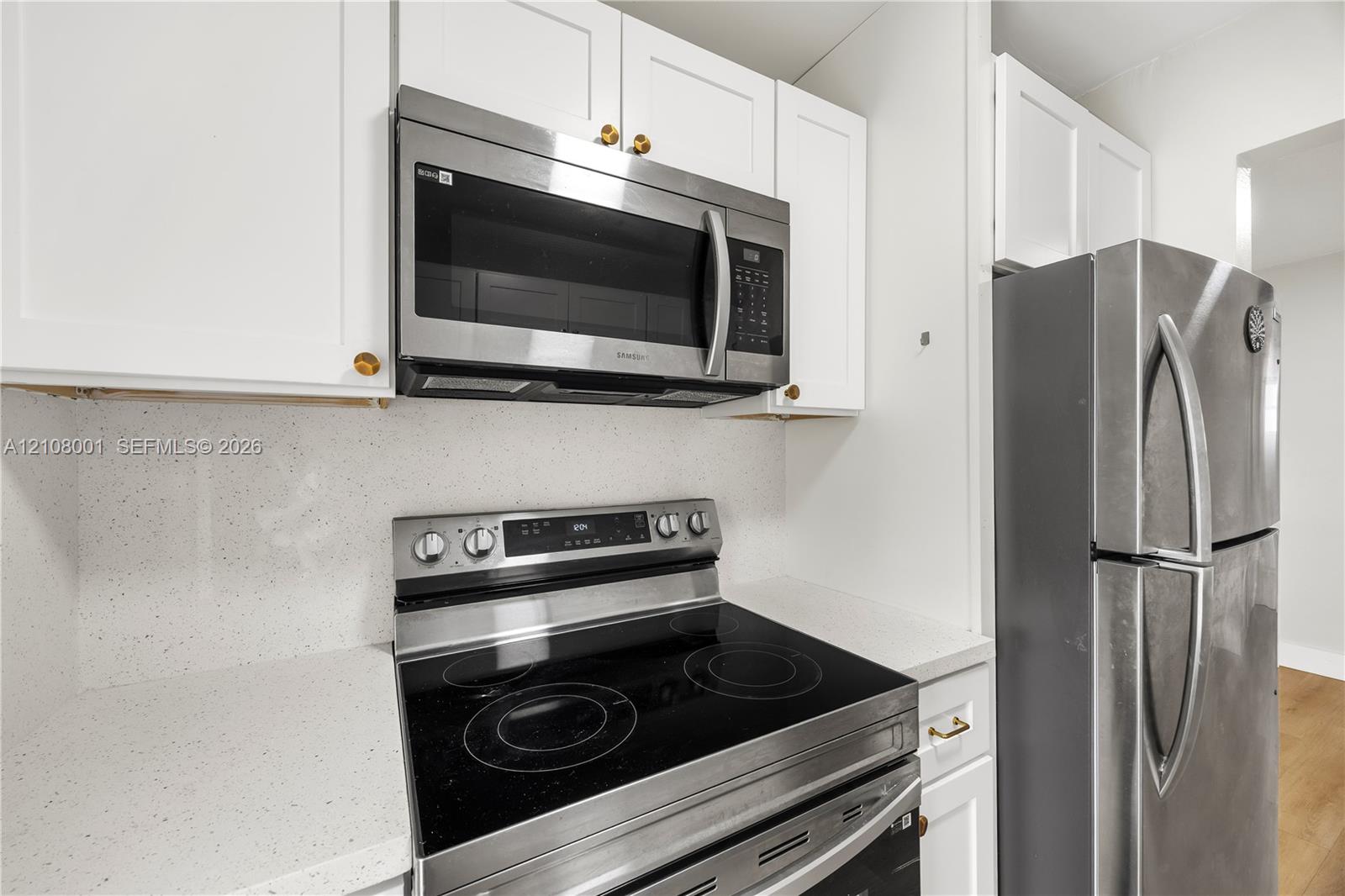White shaker-style cabinetry, stainless steel microwave, glass-top electric range, stainless steel refrigerator, and solid-surface countertop with matching backsplash