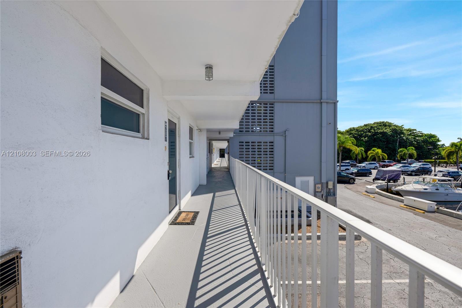 Exterior walkway featuring white stucco walls, painted concrete flooring, and a white metal railing overlooking a paved parking area and marina