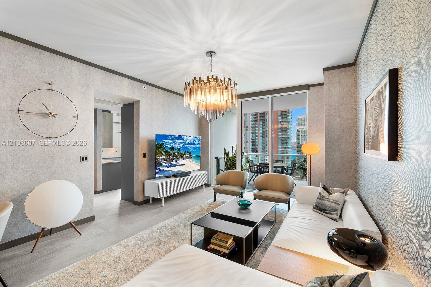 Living area featuring textured wall coverings, multi-tiered glass crystal chandelier, large-format tile flooring, and floor-to-ceiling glass sliding doors opening to an exterior balcony