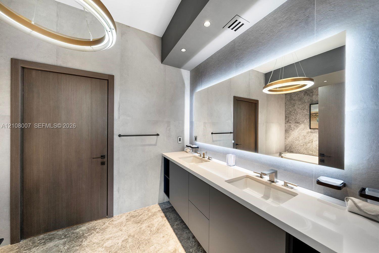 Double vanity featuring a white solid-surface countertop with integrated sinks, matte black hardware, backlit wall-to-wall mirror, and textured wall tile