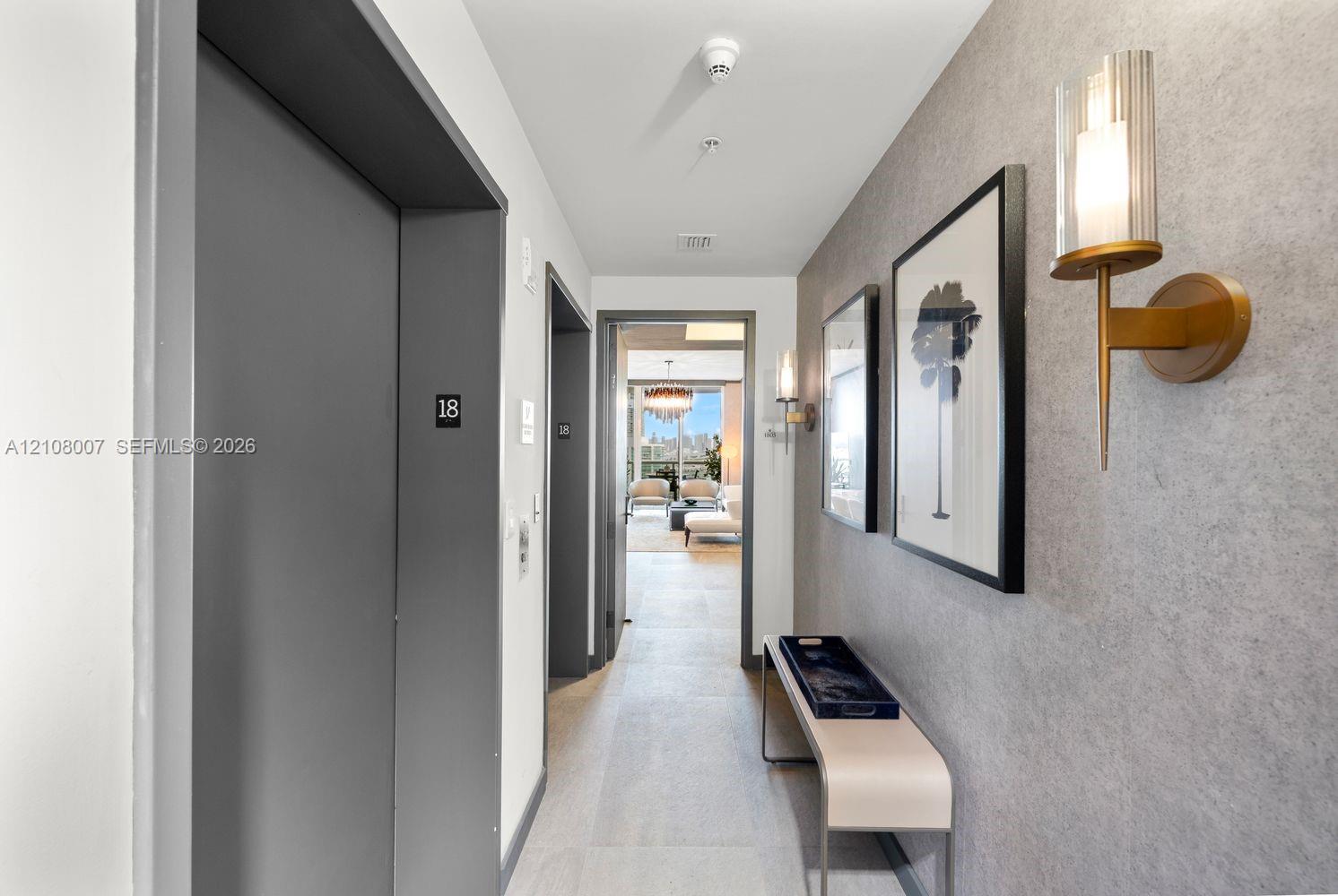 Hallway featuring textured grey wall cladding, gold-toned metallic wall sconces with cylindrical glass shades, and light-toned tile flooring