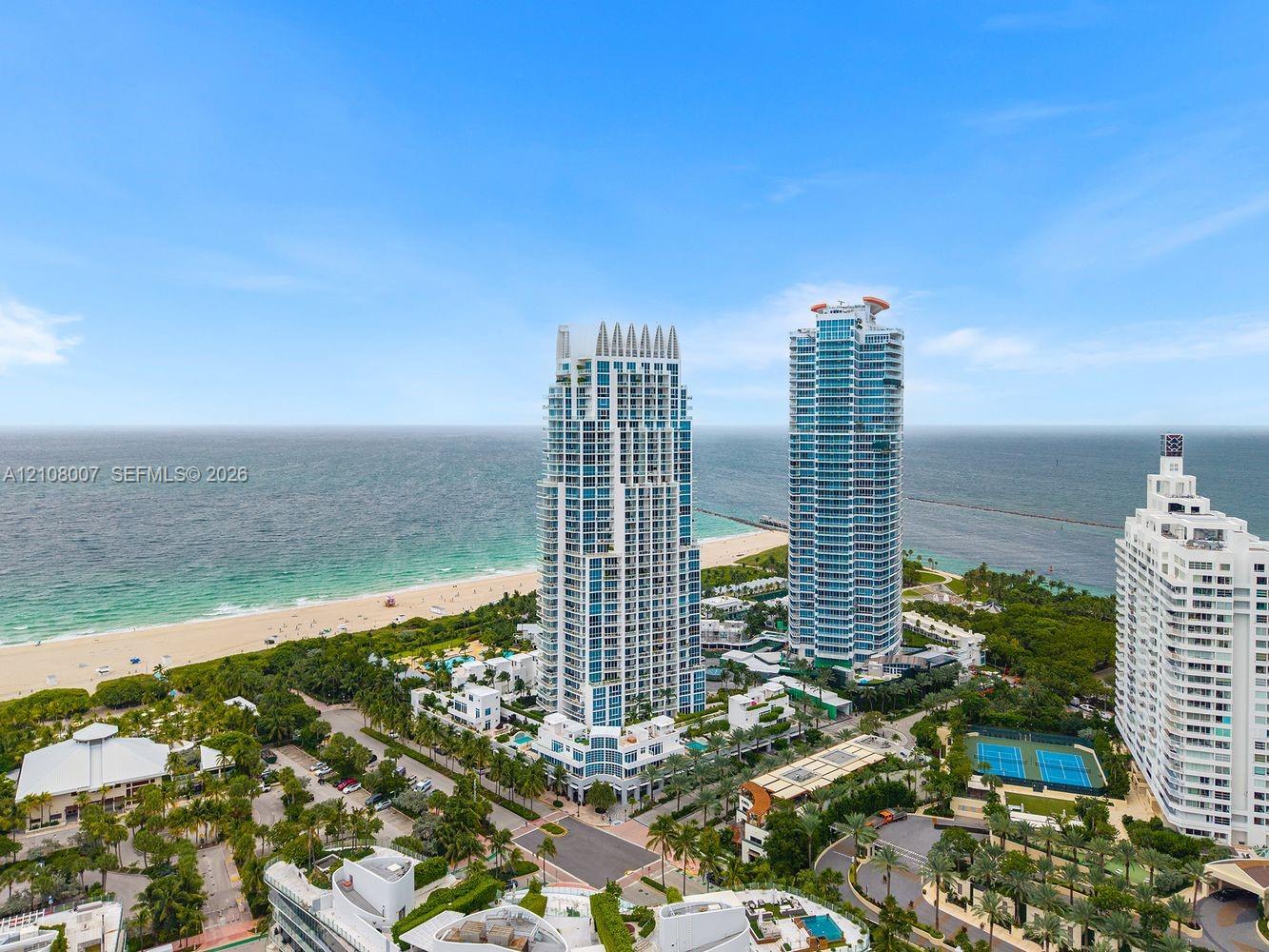 Coastal high-rise buildings situated along a sandy shoreline with direct access to the ocean, featuring manicured landscaping, paved walkways, and dual tennis courts