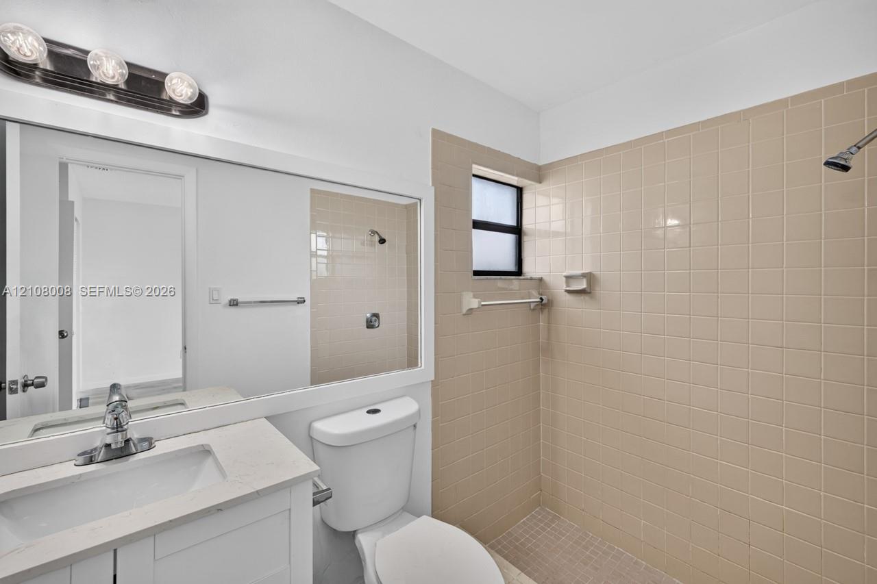 Bathroom featuring a vanity with a solid-surface countertop, rectangular integrated sink, chrome-finish faucet, wall-spanning mirror, and a tiled shower enclosure