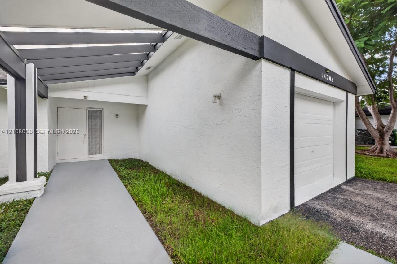 Stucco exterior featuring a covered pergola entryway, painted wood-beam detailing, a paneled garage door, and a concrete walkway