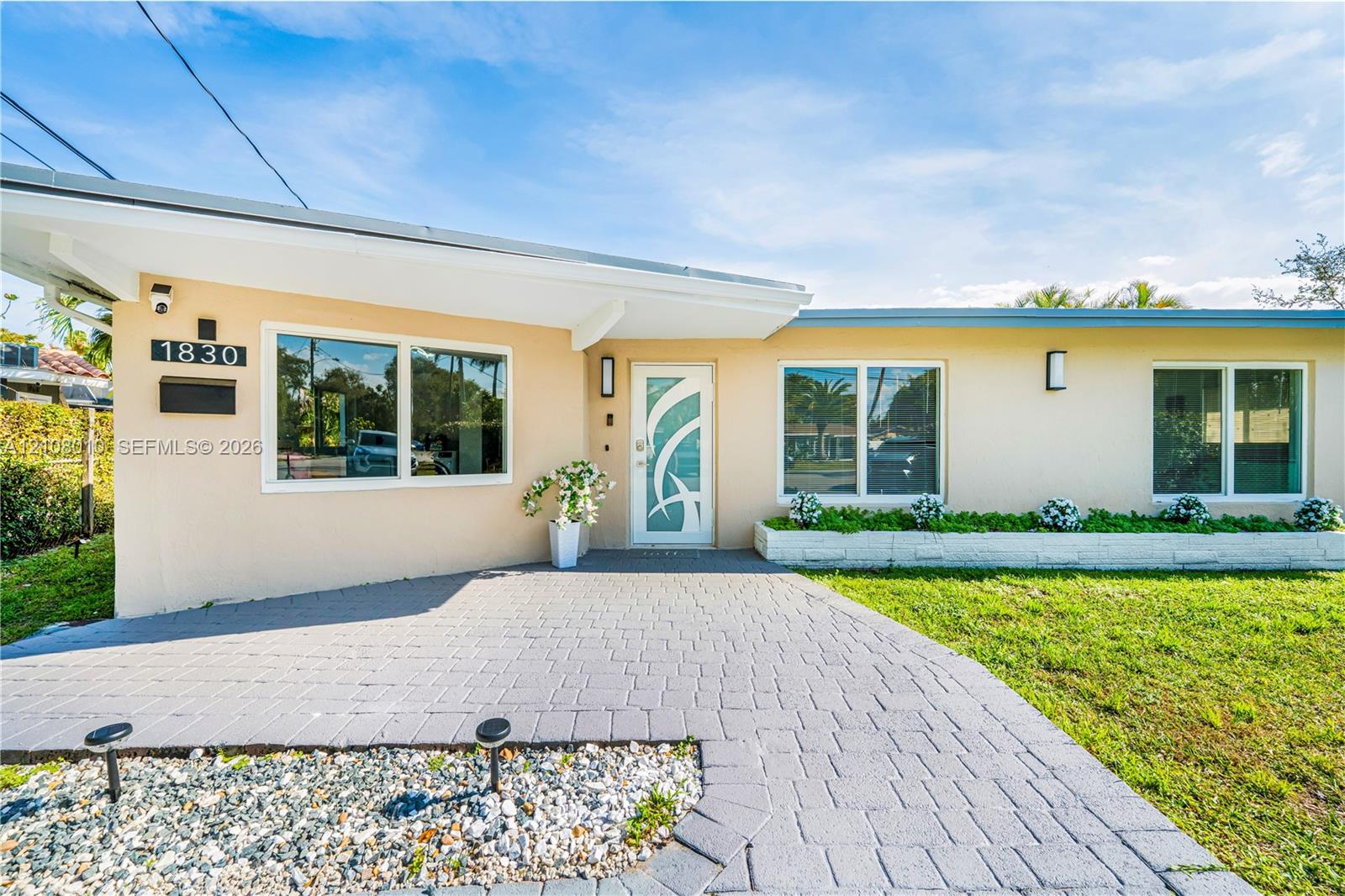 Single-story exterior featuring a paver driveway, decorative glass-paneled entry door, low-profile roofline, and integrated stone-faced planter bed