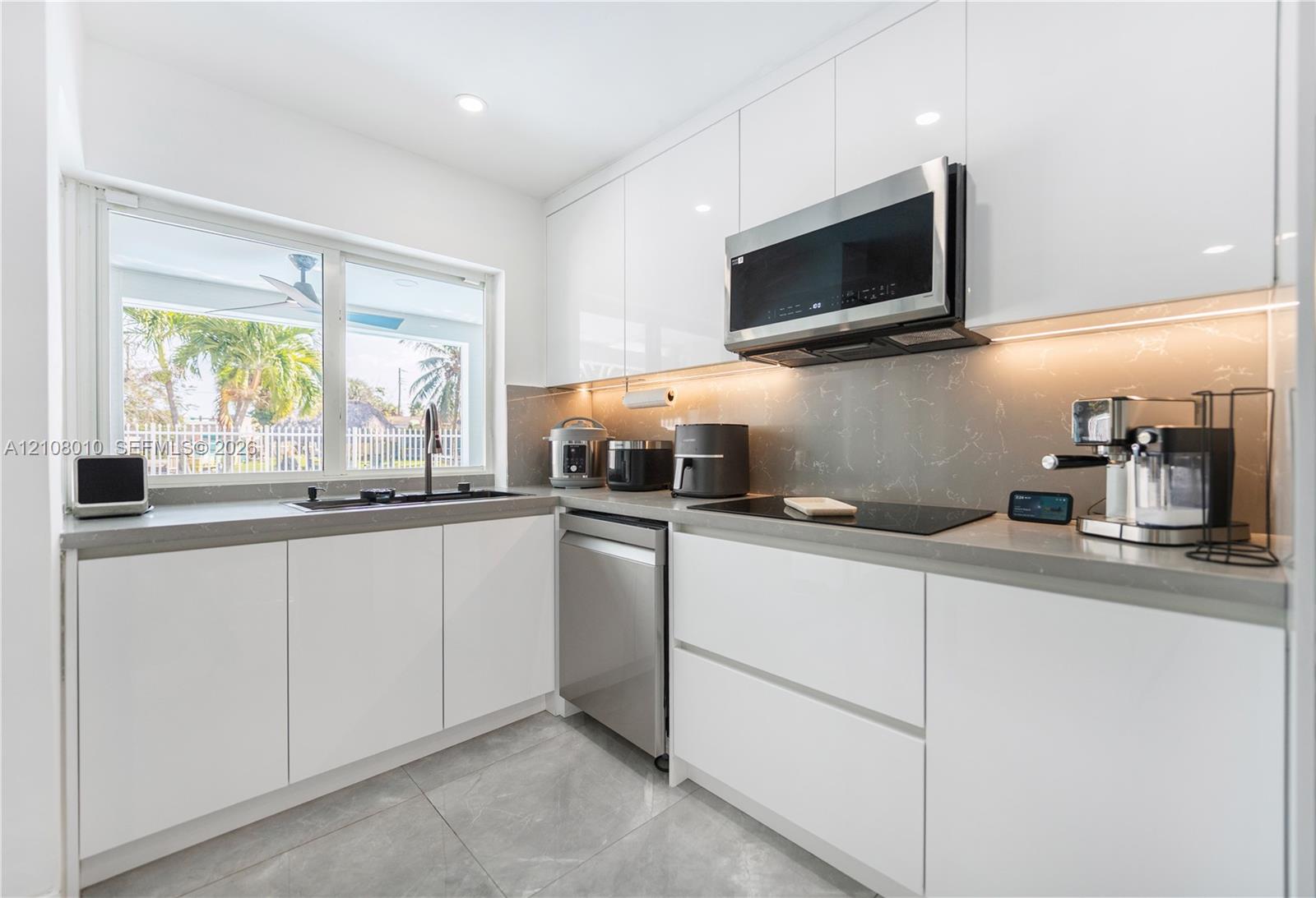 Modern kitchen featuring high-gloss white flat-panel cabinetry, solid-surface countertops, integrated under-cabinet lighting, stainless steel appliances, and large-format porcelain tile flooring