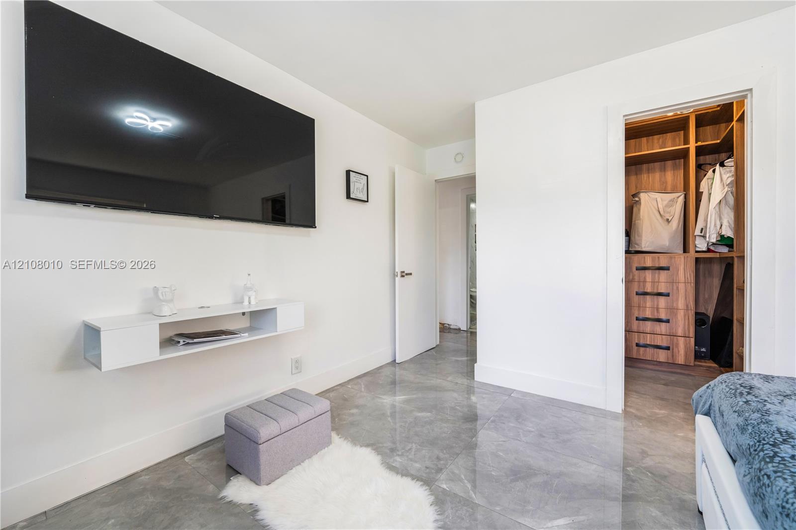 Large-format grey tile flooring, wall-mounted white floating shelf, and walk-in closet featuring built-in wood-finish cabinetry with drawers and hanging storage