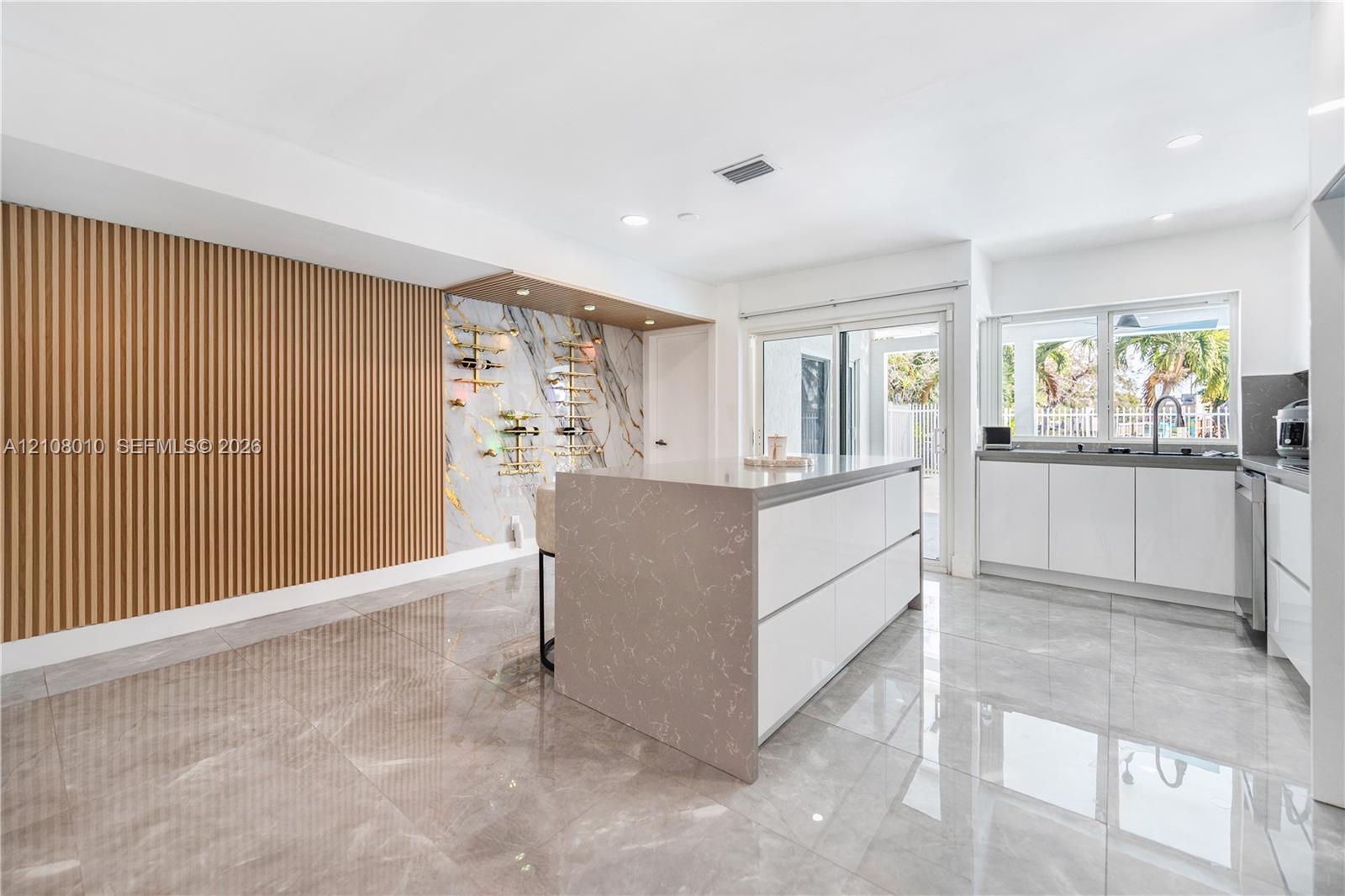 Modern kitchen featuring large-format tile flooring, a center island with solid-surface countertop, vertical wood-slat accent wall, and a custom built-in wine display with stone-finish backsplash