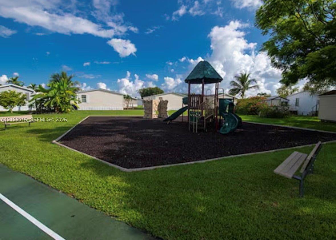 Community recreational area featuring a playground structure with a climbing wall and slide, wood-chip ground cover, and fixed benches situated on a manicured lawn