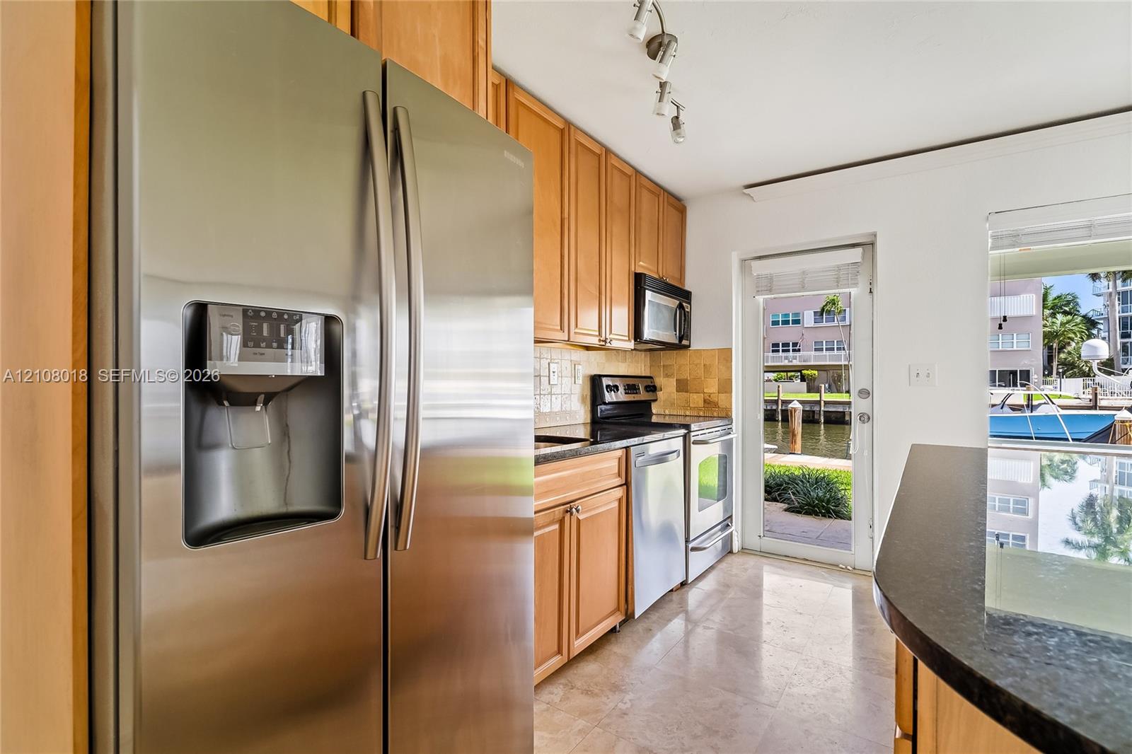 Kitchen featuring natural wood cabinetry, stainless steel appliances, dark stone-finish countertops, tile backsplash, and polished stone flooring