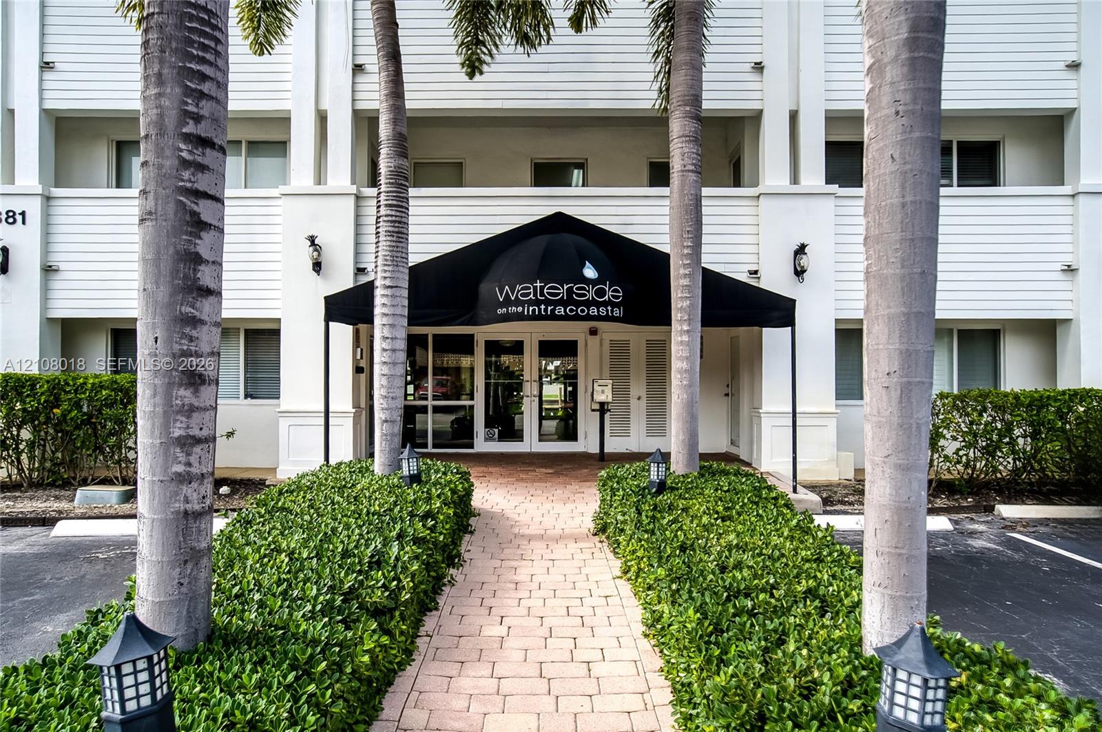 Building exterior featuring white siding, a covered entryway with a black awning, brick-paver walkway, and mature landscaping