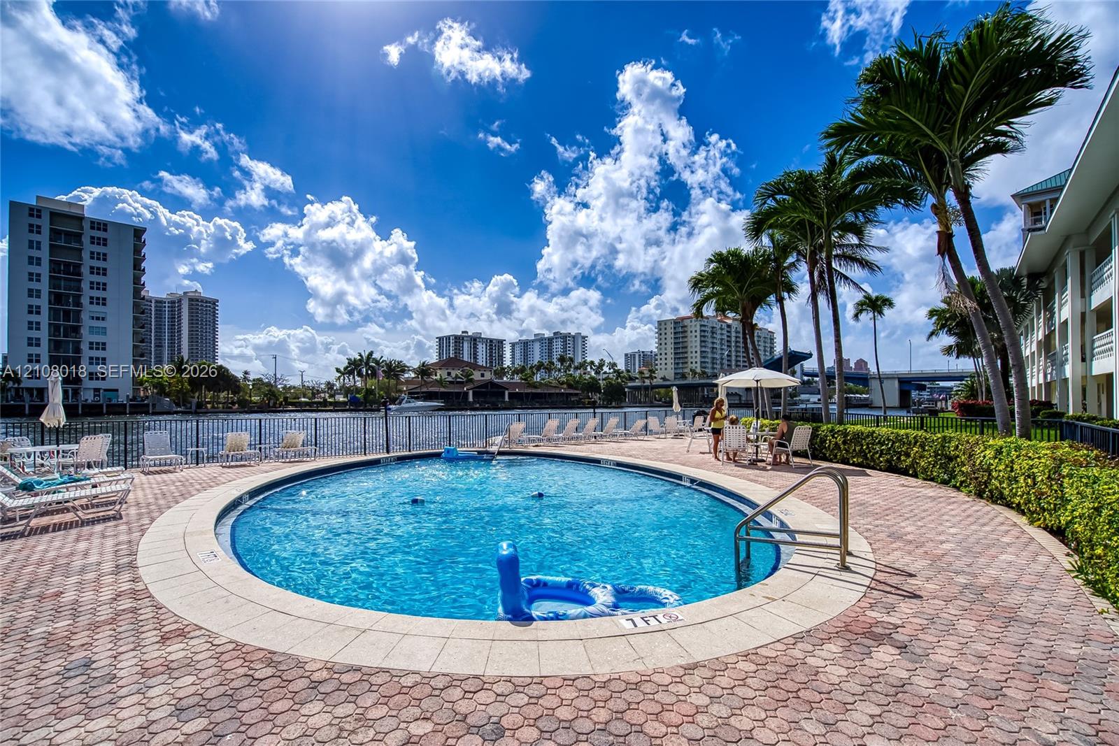 Circular swimming pool featuring stone paver decking, metal perimeter fencing, and mature palm trees