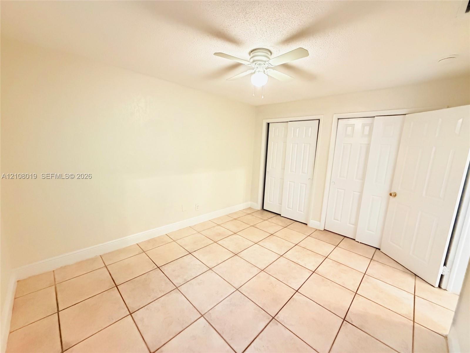 Neutral-toned ceramic tile flooring, dual sets of white closet doors, and a ceiling fan with integrated lighting