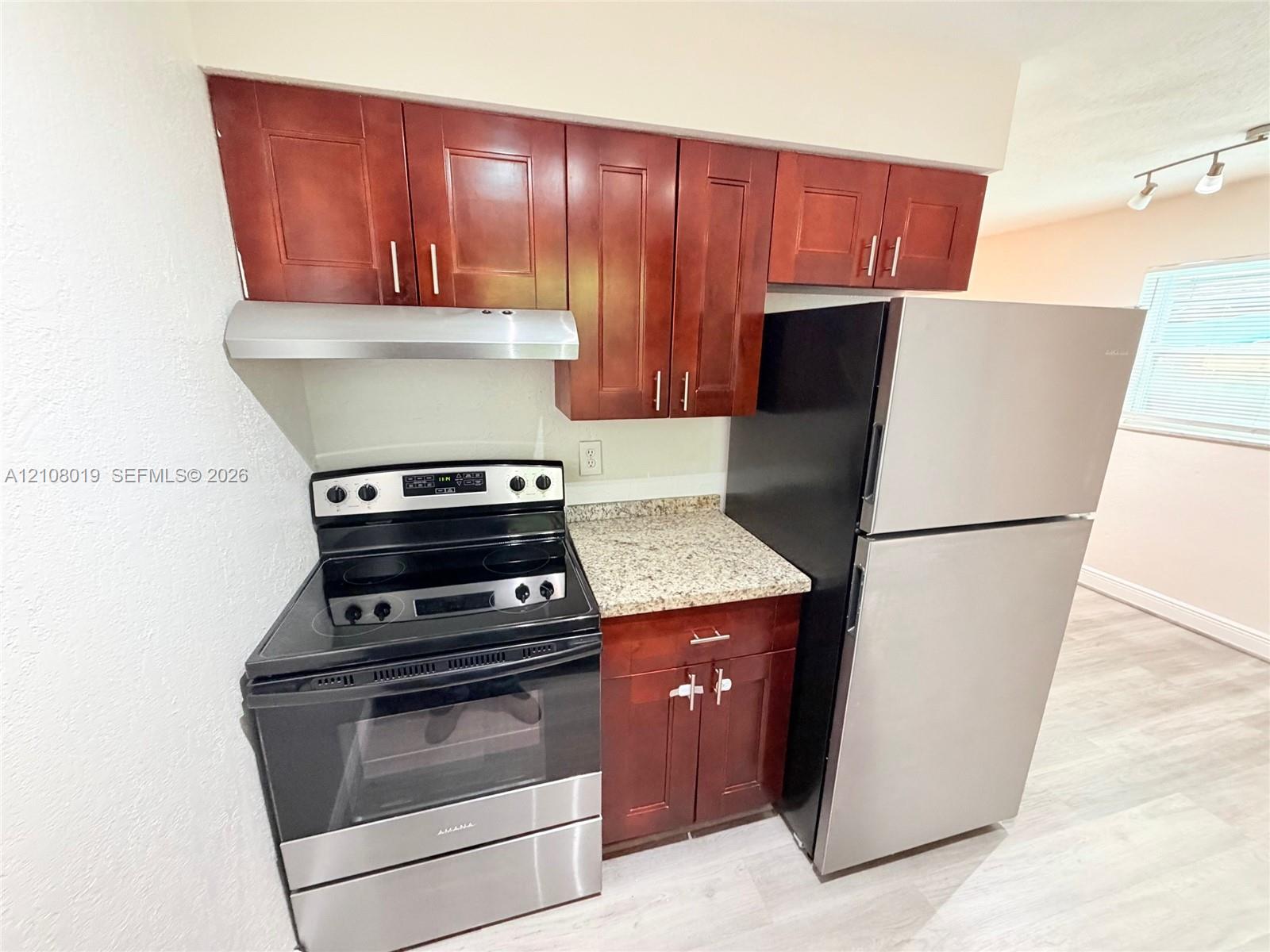 Kitchen area featuring wood-finish cabinetry, stone-patterned countertops, a stainless steel electric range, a stainless steel range hood, and a stainless steel refrigerator