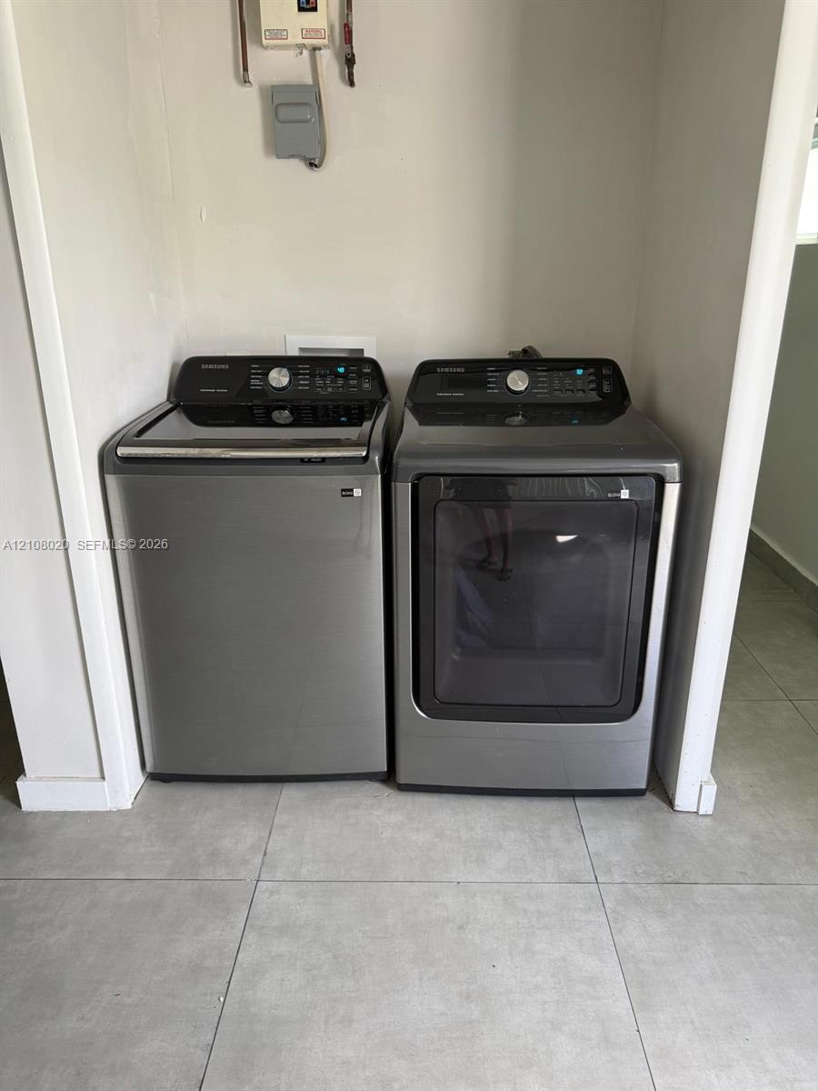 Dedicated laundry alcove featuring large-format ceramic tile flooring and charcoal-finished top-load washing machine and front-load dryer
