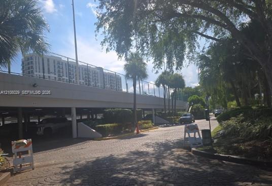 Covered parking structure featuring stone-paver driveway, concrete canopy, chain-link perimeter fencing, and mature palm landscaping