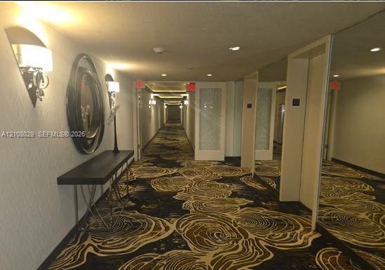 Hallway featuring patterned wall-to-wall carpeting, decorative wall-mounted sconces, textured wall coverings, and mirrored paneling