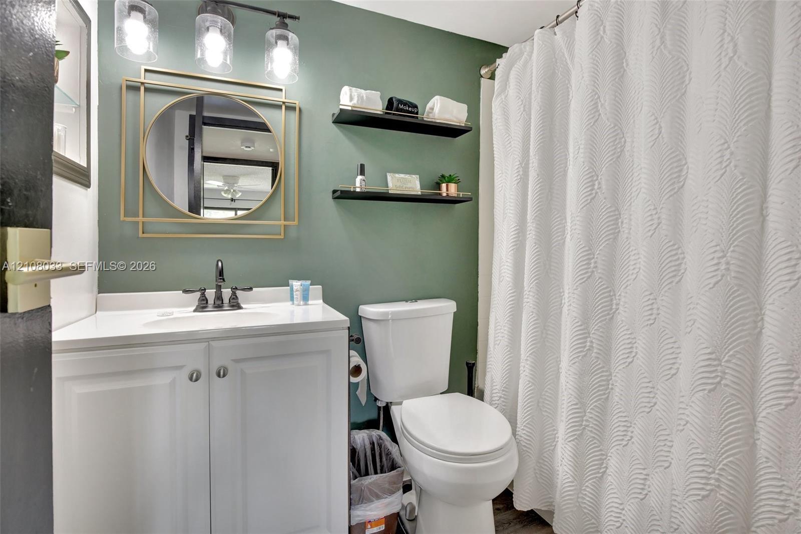 Bathroom featuring a white vanity cabinet, solid-surface countertop with an integrated basin, three-light fixture with glass shades, geometric gold-tone wall mirror, and dual wall-mounted floating shelves