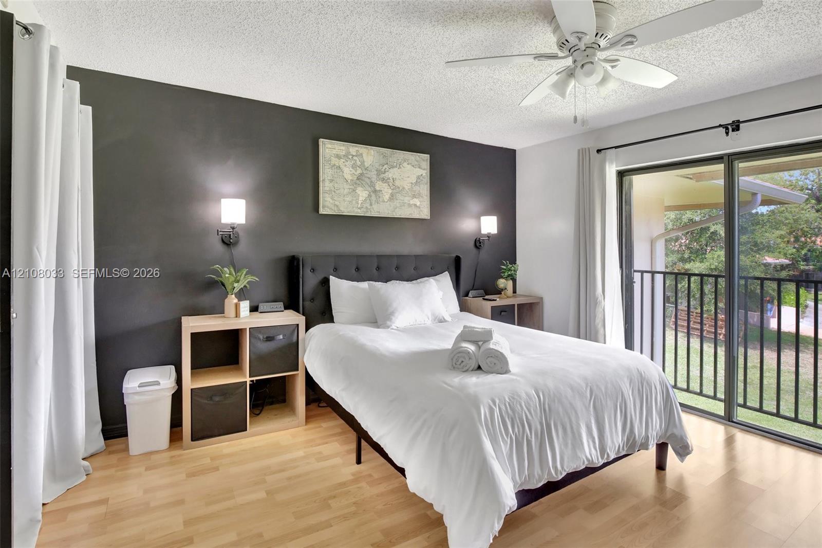 Bedroom featuring wood-finish flooring, a dark accent wall, two wall-mounted light fixtures, a ceiling fan, and sliding glass doors leading to a balcony