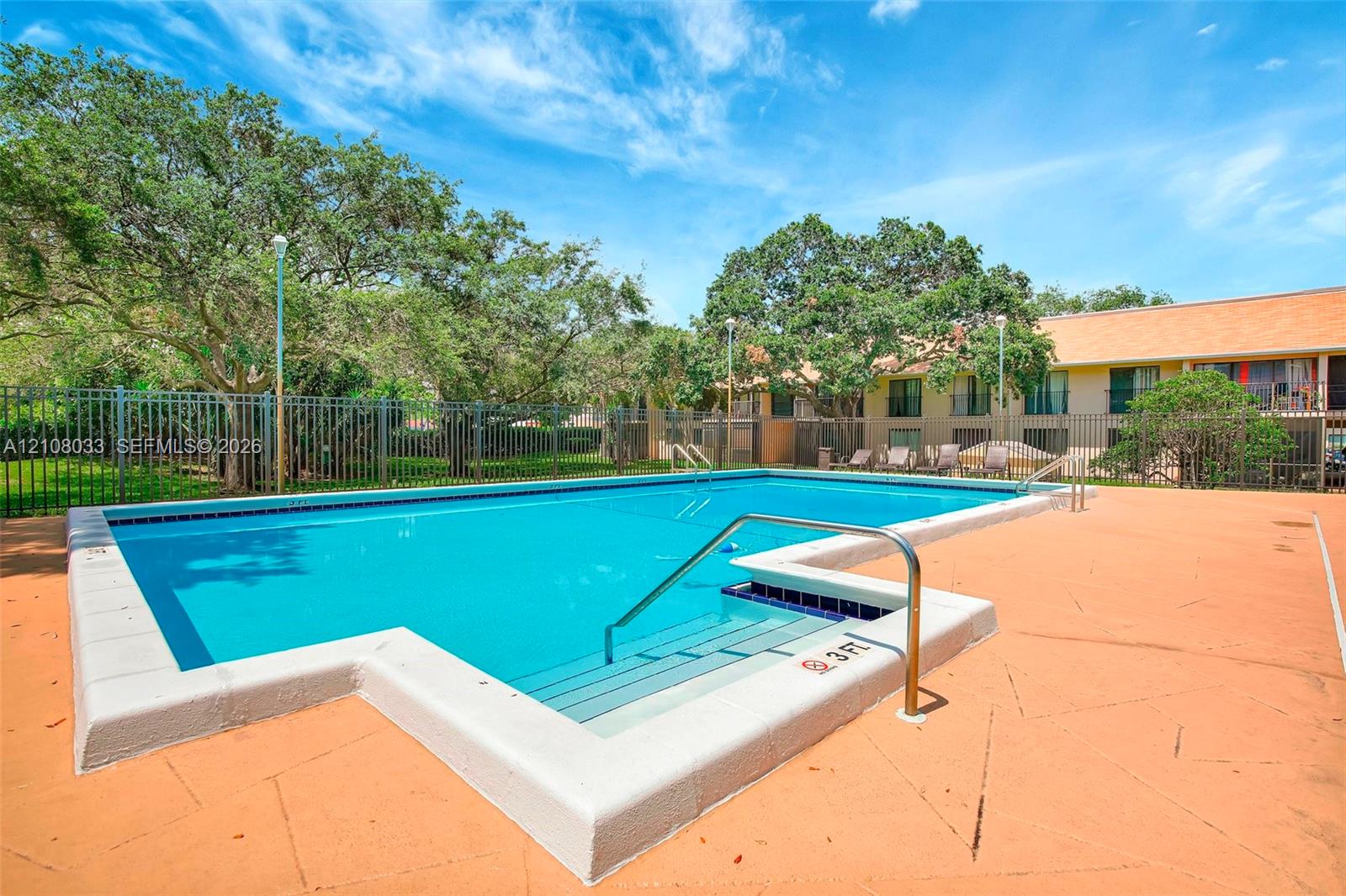 Rectangular swimming pool featuring a concrete deck, metal perimeter fencing, integrated steps with a metal handrail, and mature trees