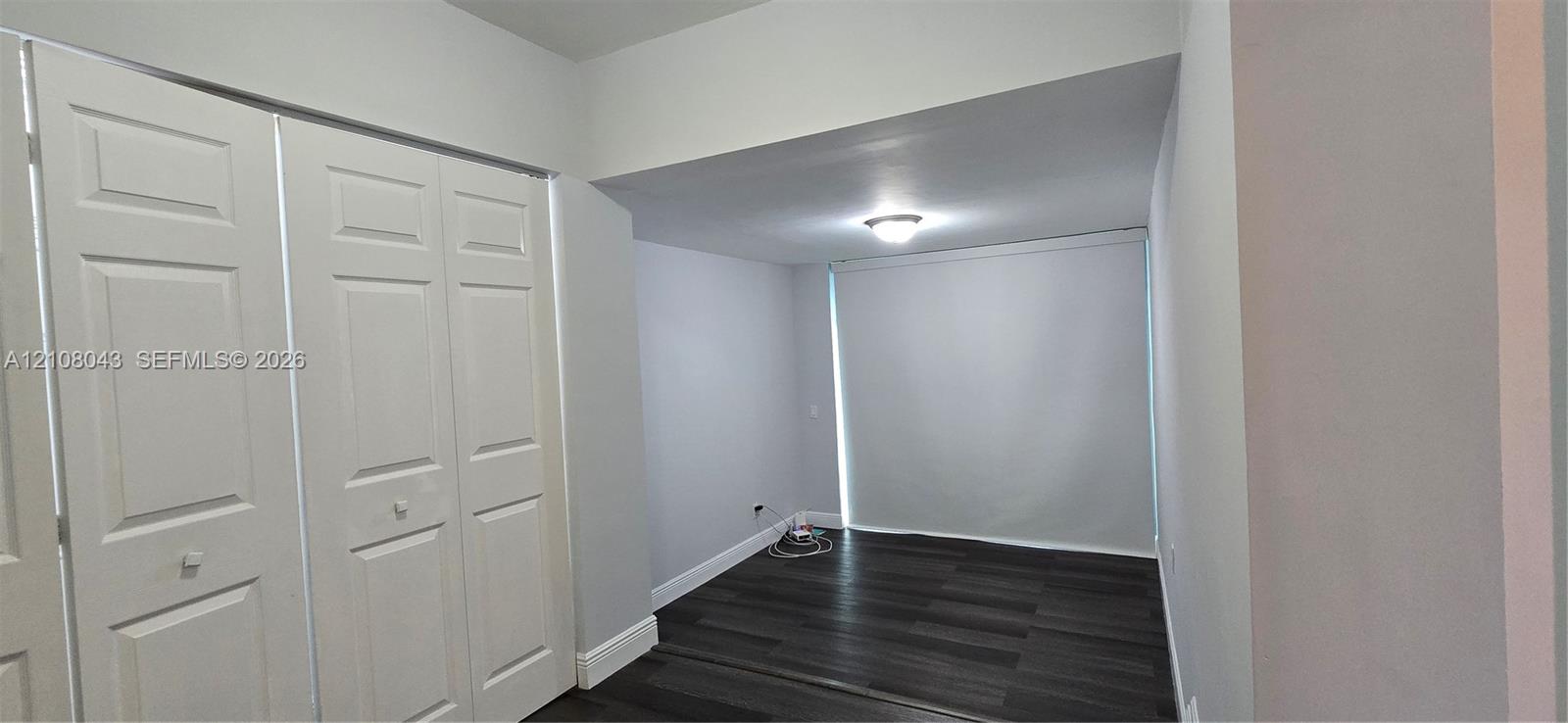 Room interior featuring dark wood-finish flooring, white panel closet doors, neutral wall paint, and a flush-mount ceiling light fixture