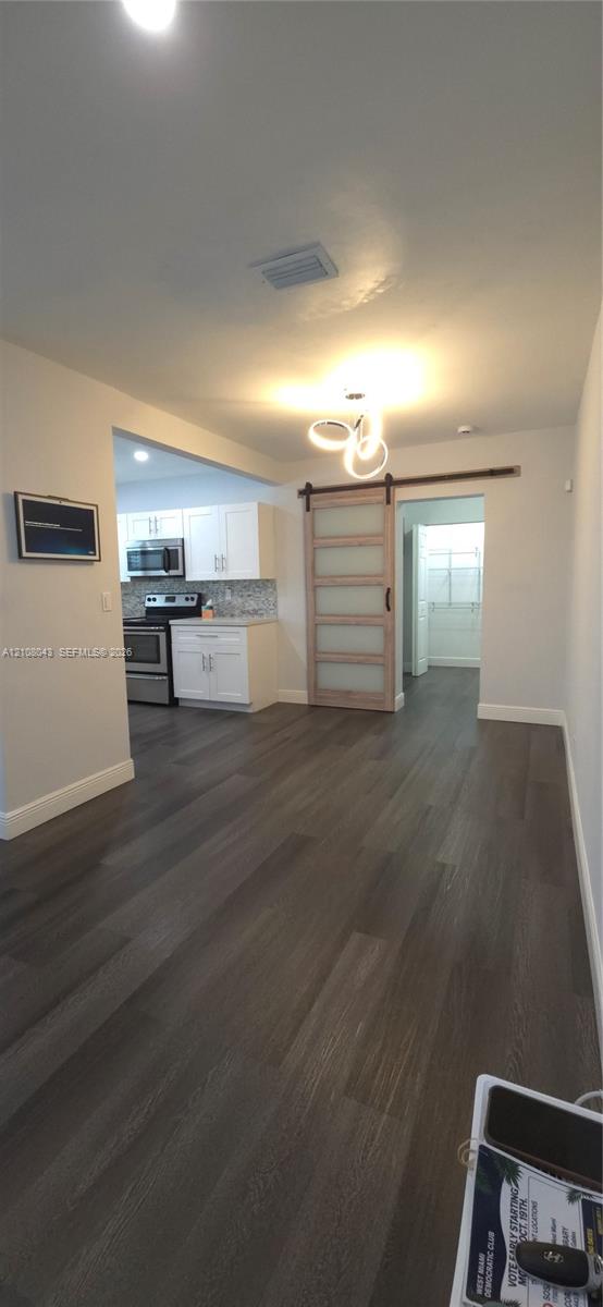 Wood-finish flooring, sliding glass-panel barn door with metal hardware, contemporary three-ring light fixture, white kitchen cabinetry, and stainless steel kitchen appliances