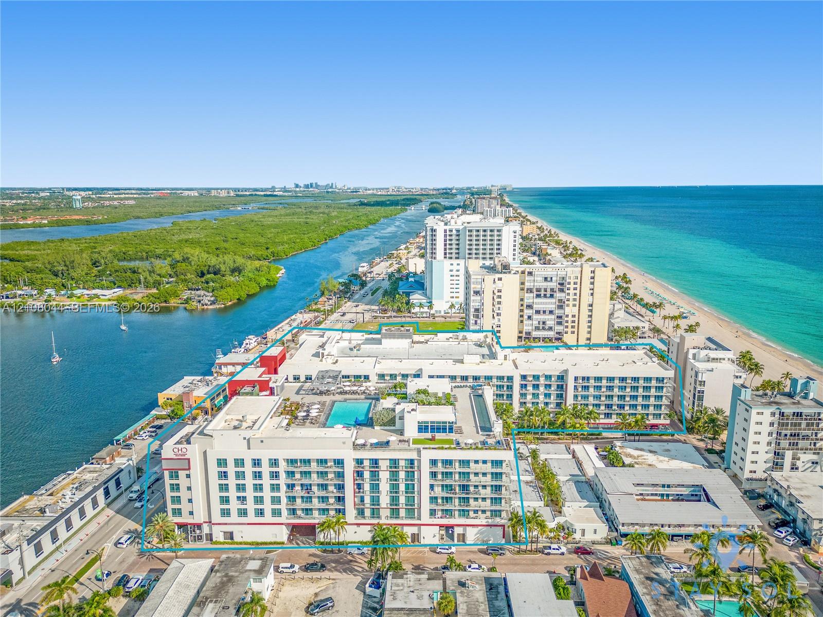 Aerial view of a coastal multi-story residential structure featuring a rooftop pool, private balconies, and proximity to both the oceanfront and a bay-side waterway