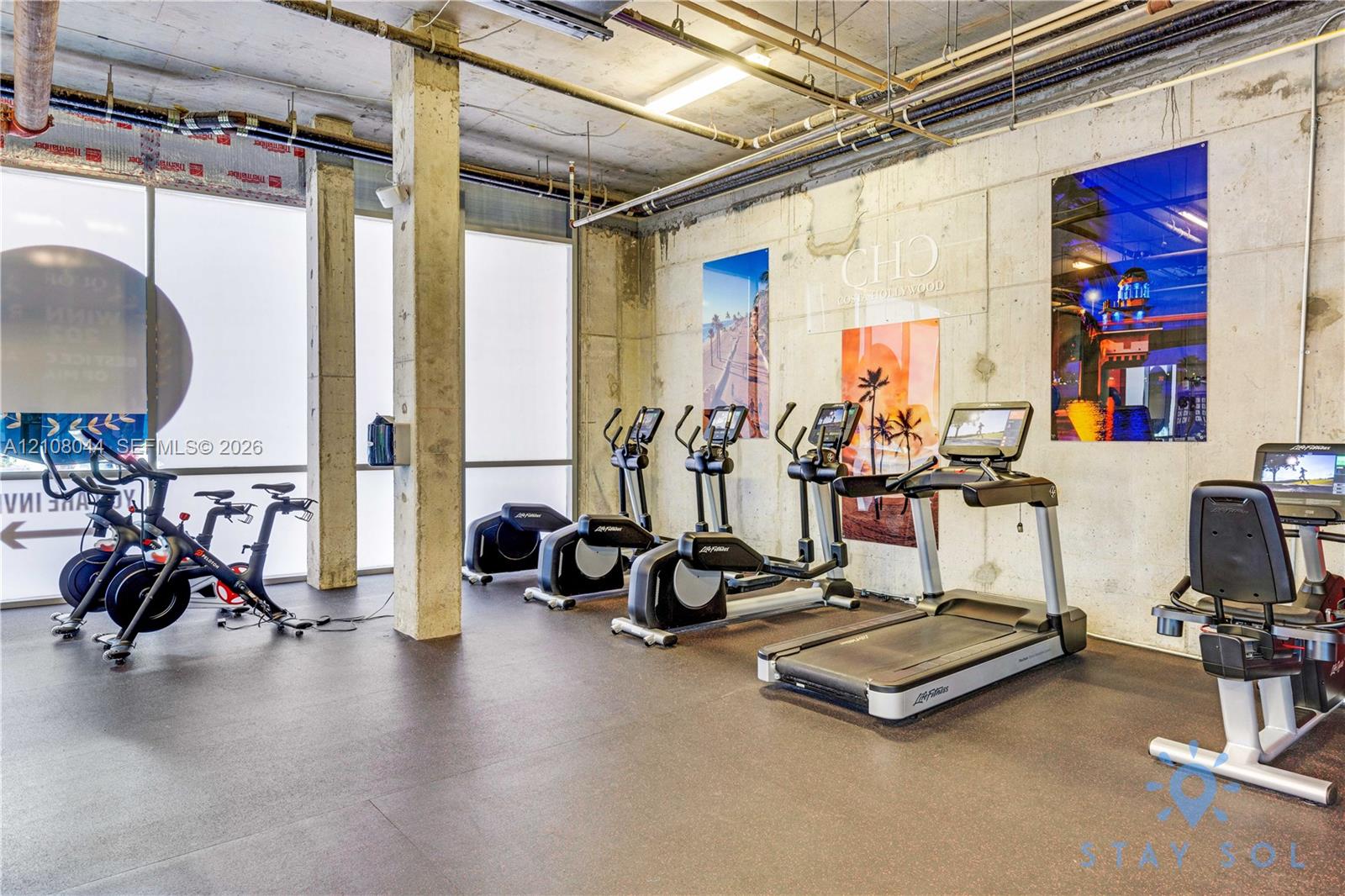 Fitness room featuring exposed concrete walls and ceiling, industrial-style piping, floor-to-ceiling windows, and rubber flooring
