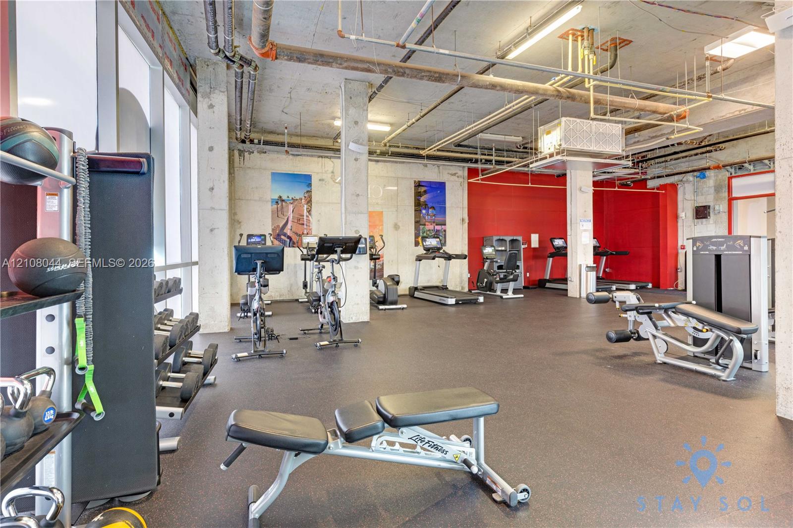 Industrial-style fitness center featuring high exposed ceilings, concrete structural columns, rubberized flooring, and expansive glass window panels