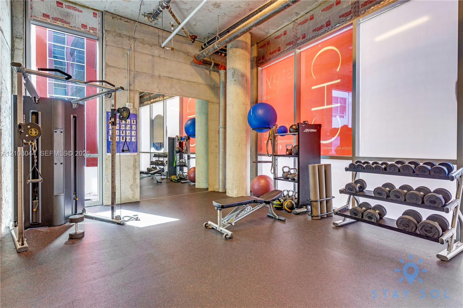 Industrial-style fitness space featuring exposed concrete ceilings, floor-to-ceiling windows, rubberized flooring, and an exposed structural pillar