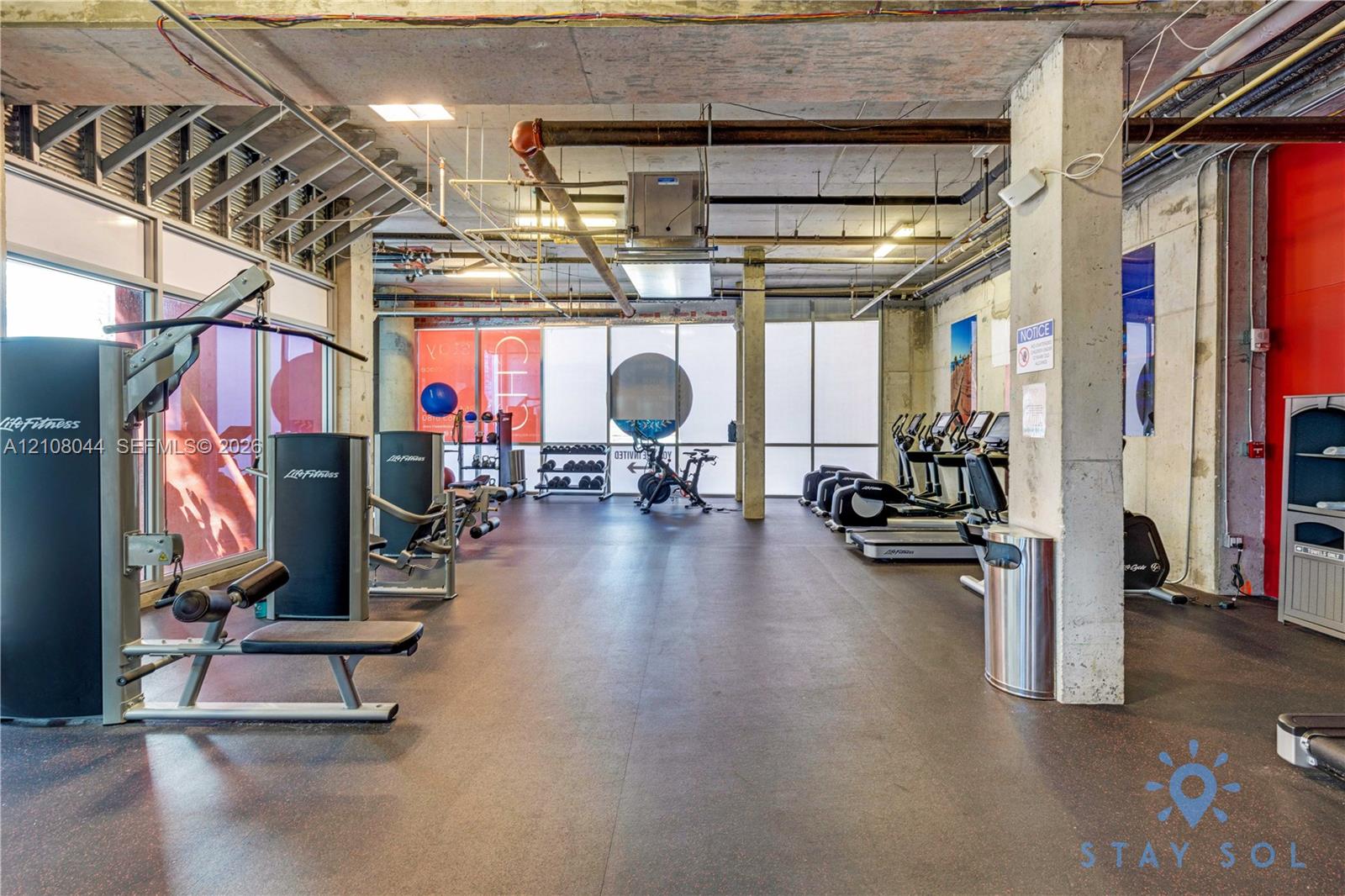 Industrial-style exercise facility featuring exposed concrete ceilings, large-scale floor-to-ceiling windows, rubberized flooring, and structural concrete columns