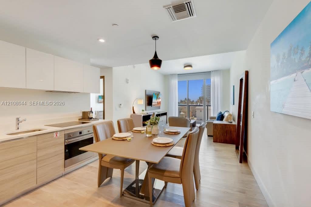 Open-concept area featuring a sleek kitchen with high-gloss white cabinetry, light wood-finish lower cabinetry, an integrated stainless steel oven, and light-toned flooring, highlighted by floor-to-ceiling glass paneling