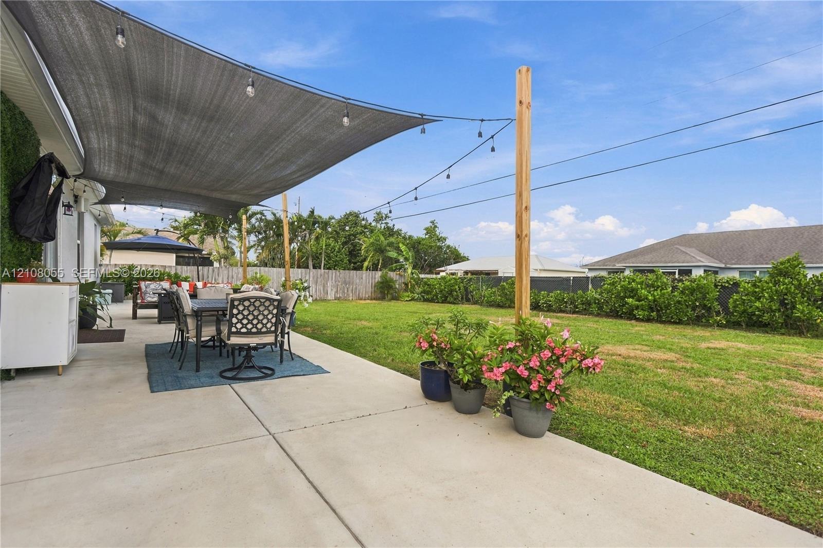 Concrete patio featuring a retractable shade sail, overhead string lighting, and wood-post support, extending into a fenced backyard with perimeter greenery