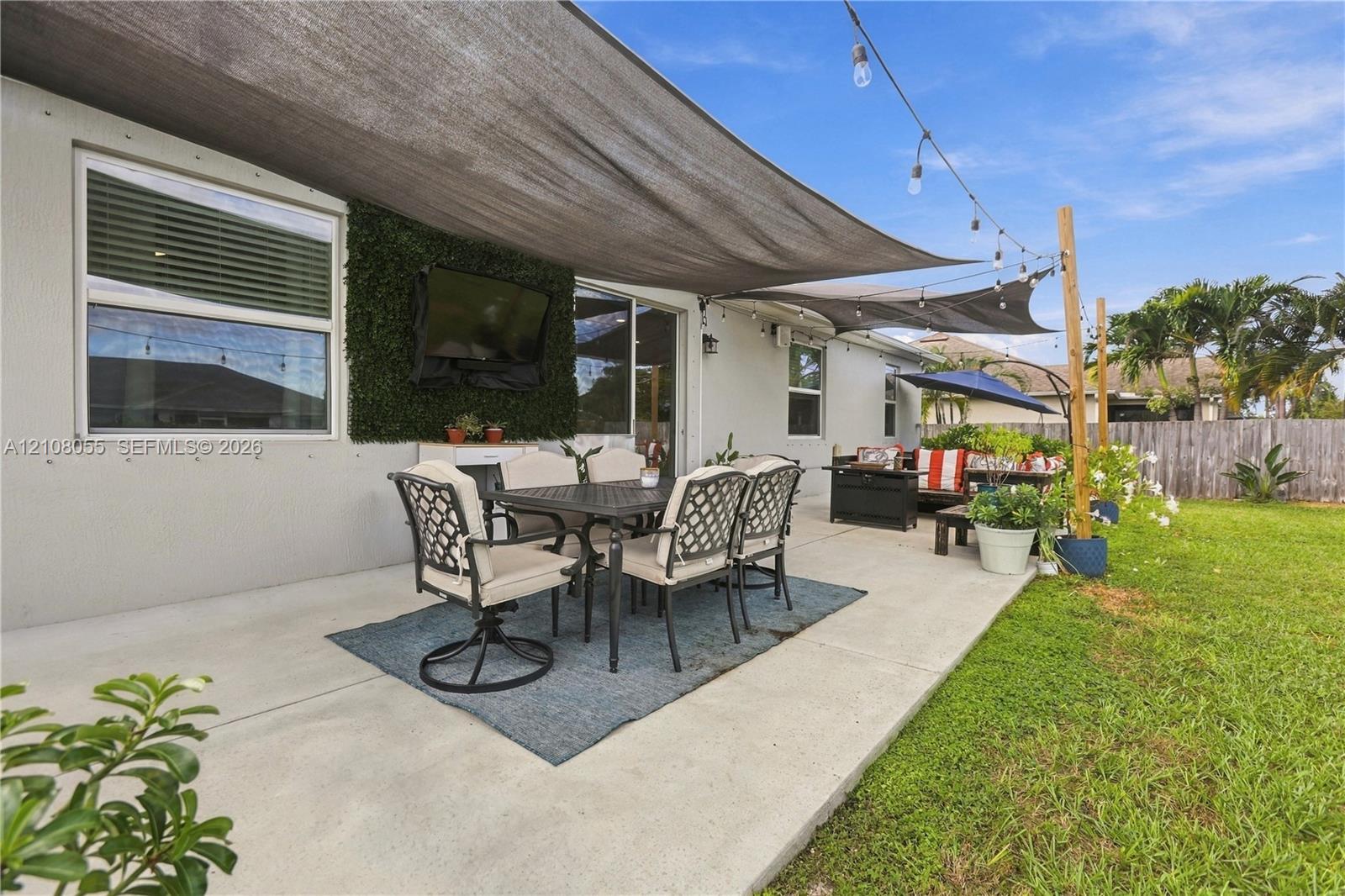 Covered concrete patio featuring tension shade sails, exterior string lighting, artificial hedge wall accent, and a fenced backyard